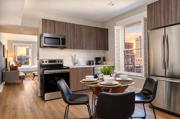 A modern kitchen with a built-in oven, microwave, and stainless steel refrigerator. There is a dining area with a round table and chairs, and a view of the city through the window. - The Penn Hotel - Smart TV, King Bed & Walkability! (San Diego)