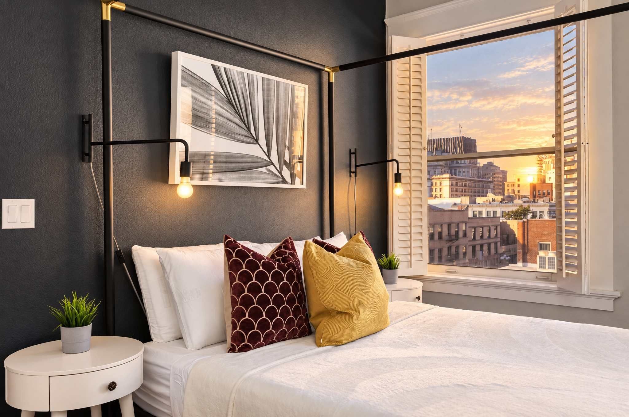 A modern bedroom with a large bed, a city view through the window, and a round nightstand with a plant.