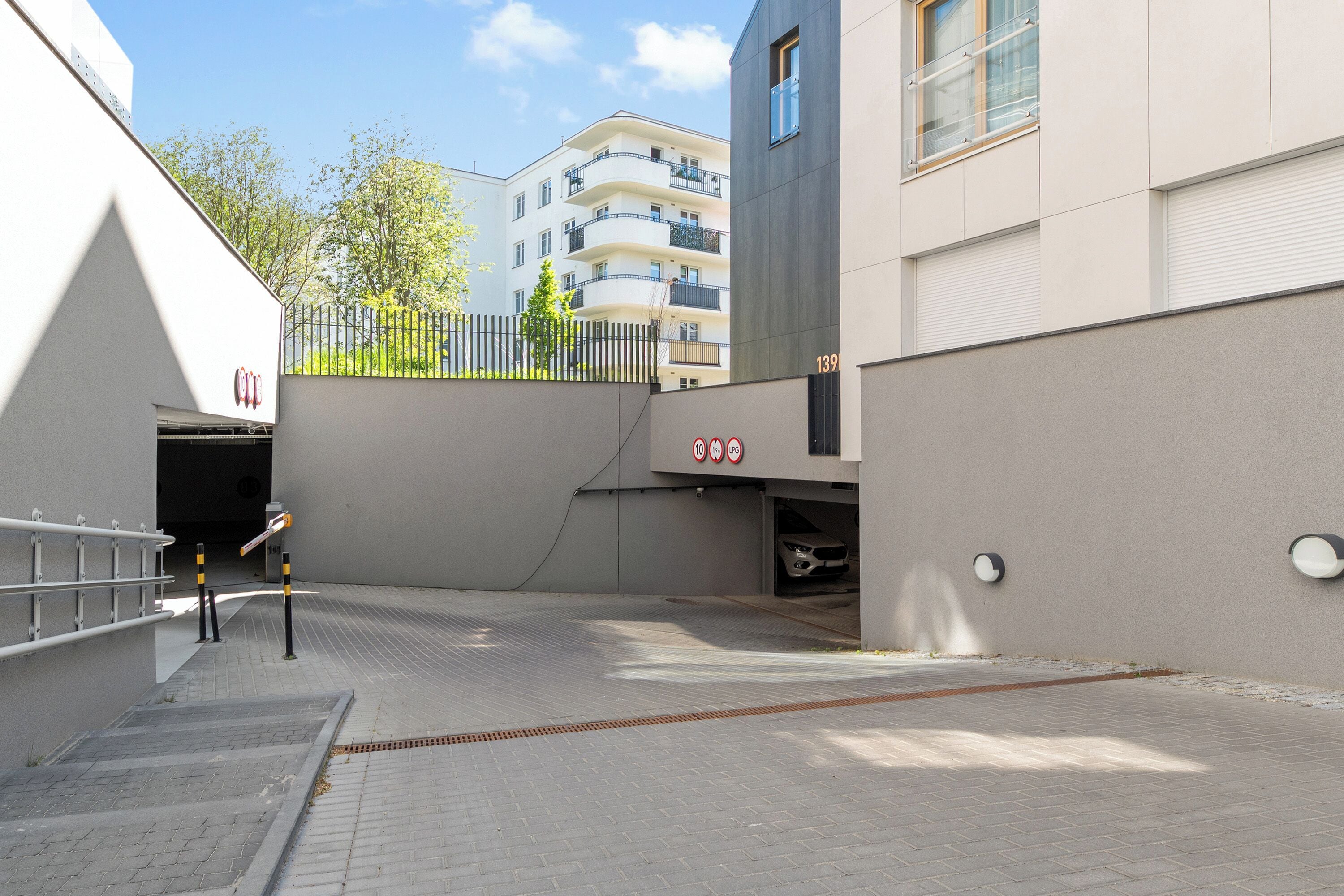 L’entrée d’un parking, avec une rampe menant à une porte de garage, une clôture en métal et un bâtiment avec un balcon en arrière-plan.