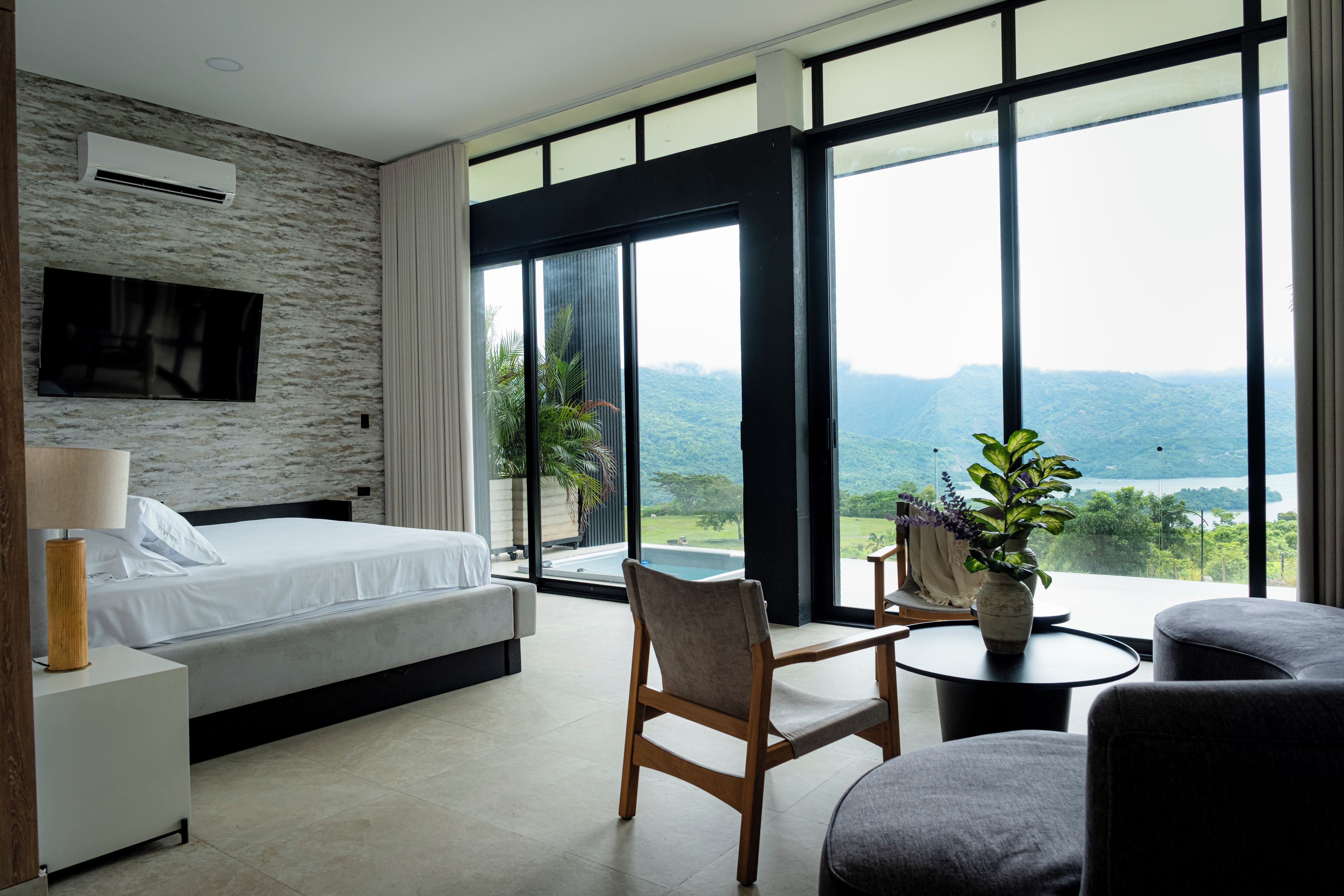 A modern bedroom with a bed, a chair, a small table, and a large window offering a view of a mountainous landscape.