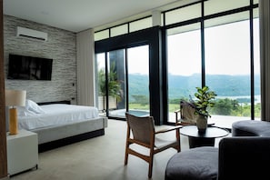 A modern bedroom with a bed, a chair, a small table, and a large window offering a view of a mountainous landscape.
