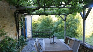 Ein Außenbereich mit Sitzgelegenheiten, einem Tisch und Bänken, umgeben von Steinmauern und viel Grün.