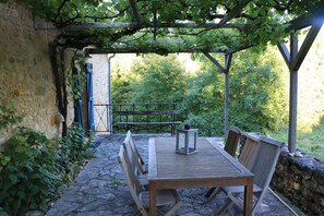 Ein Außenbereich mit Sitzgelegenheiten, einem Tisch und Bänken, umgeben von Steinmauern und viel Grün.