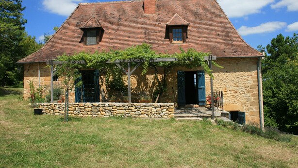 Ein Steinhaus mit Ziegeldach, zwei Fenstern und einer überwachsenen Veranda.