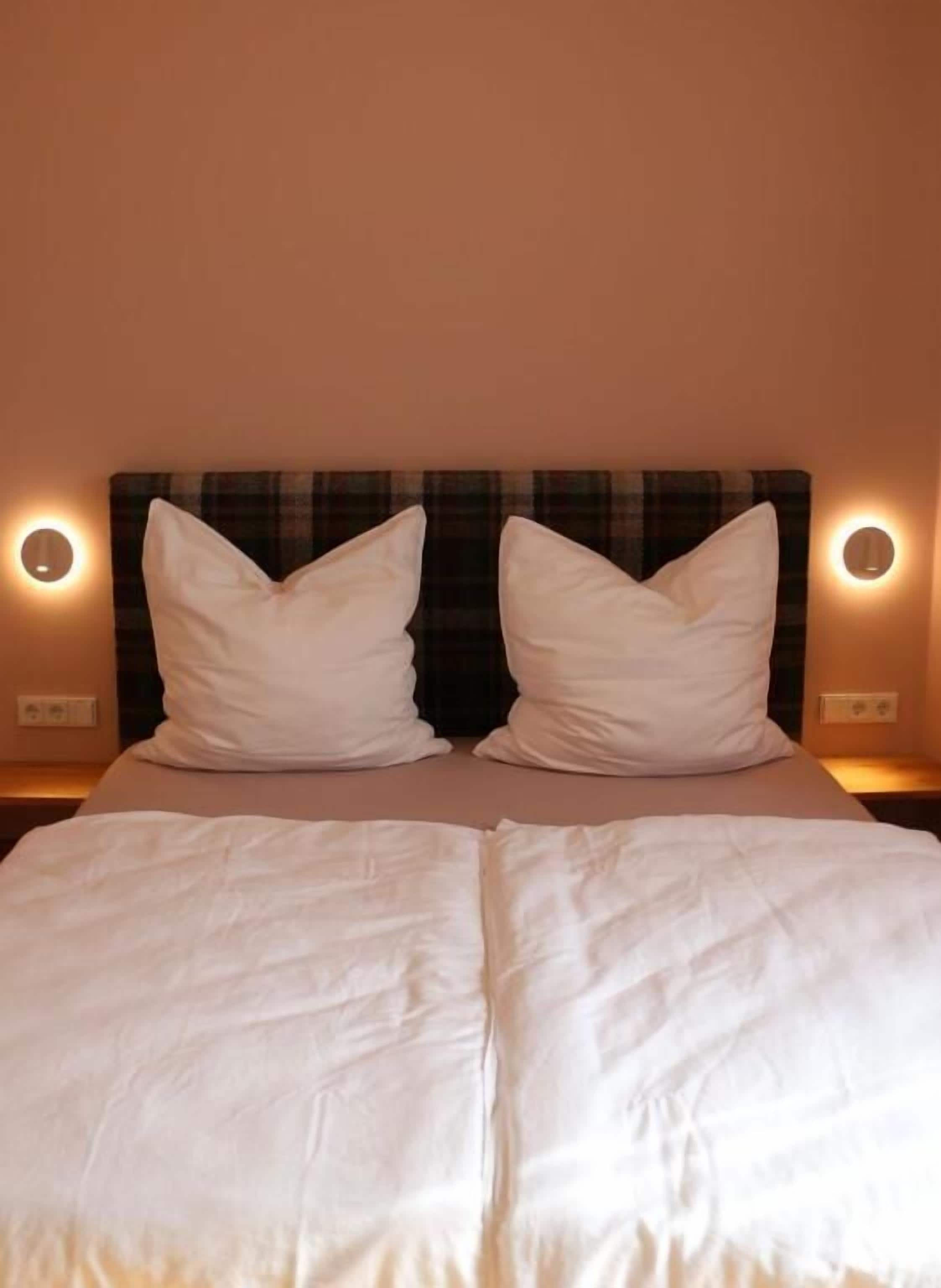 A bed with white bedding and a plaid headboard, illuminated by wall sconces.