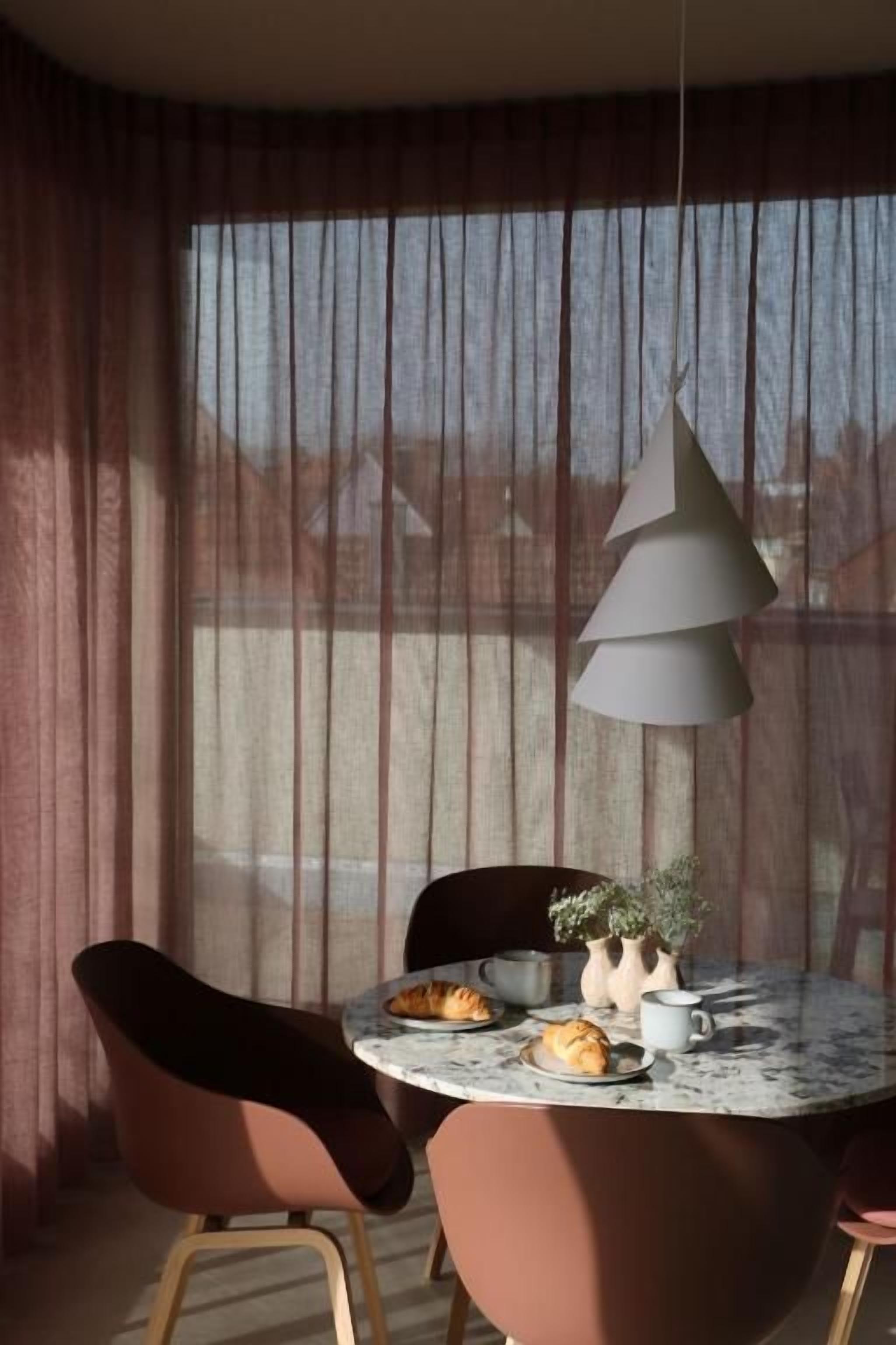 Un coin repas comprenant une table ronde, des chaises, un luminaire suspendu et une fenêtre avec des rideaux.