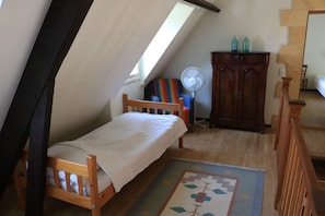 Ein kleiner, gemütlicher Raum mit schräger Decke, einem Einzelbett, einer Holzkommode und einem kleinen Ventilator.