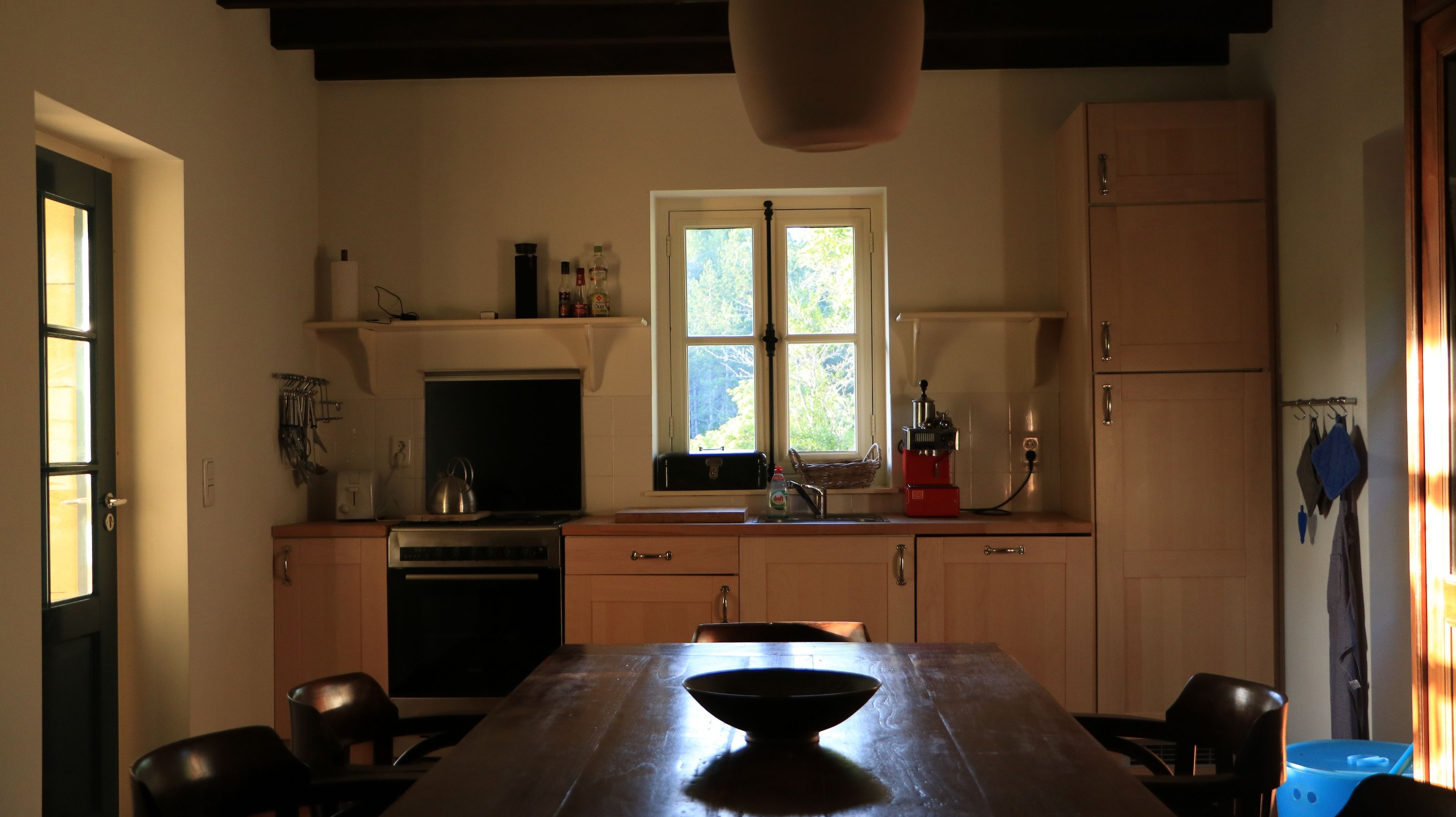 Une cuisine équipée d’une table à manger, de chaises, d’une bouilloire, d’une machine à café et d’une fenêtre donnant sur la forêt.