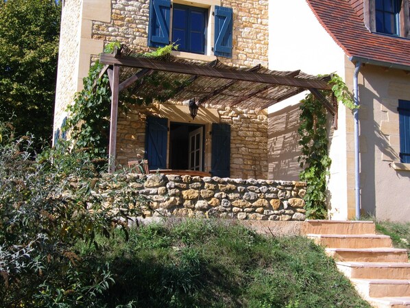 Um edifício de pedra com varanda coberta, degraus de pedra e pérgula de madeira.