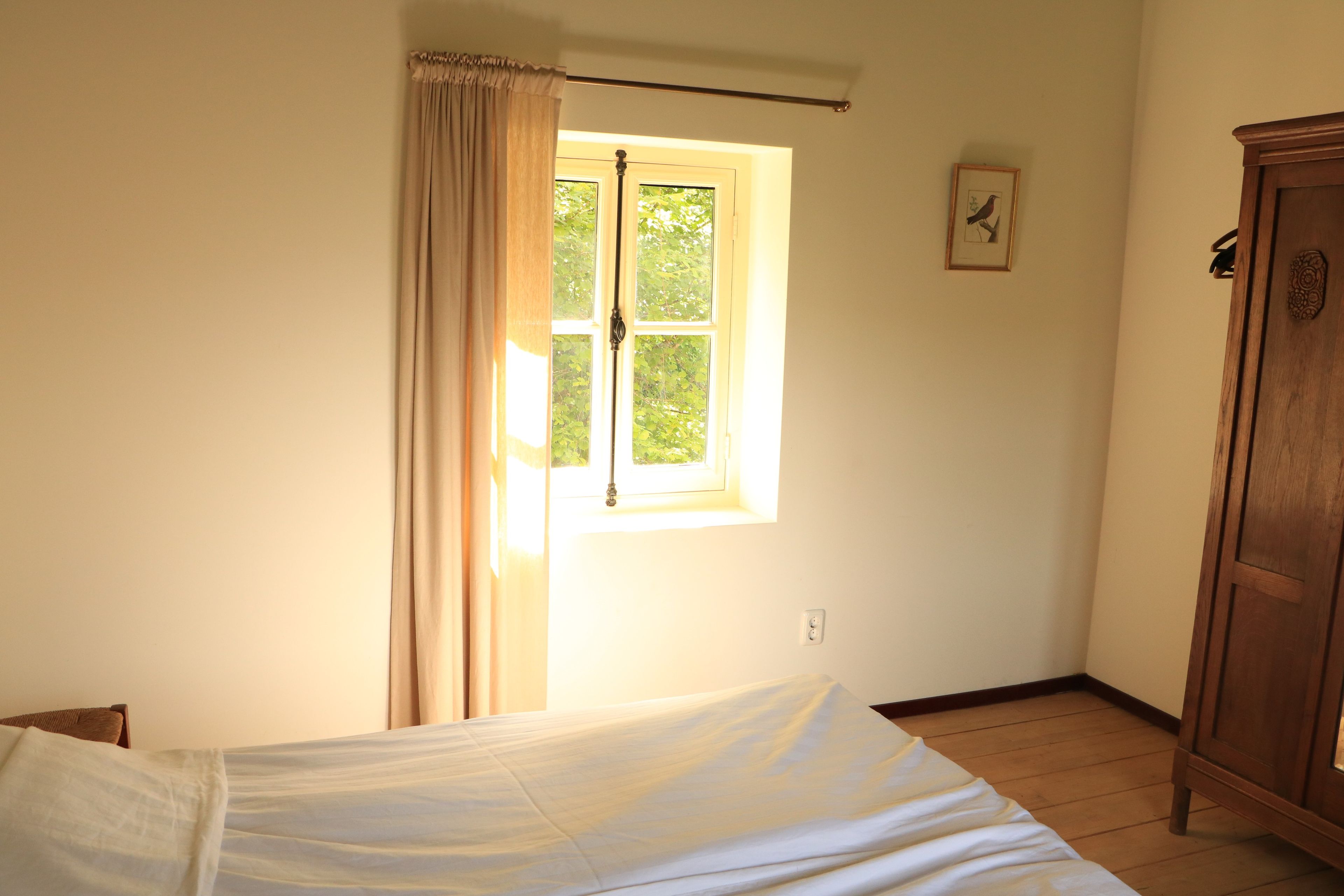 A room with a bed, a window with curtains, a wooden wardrobe, and a wall-mounted light switch.
