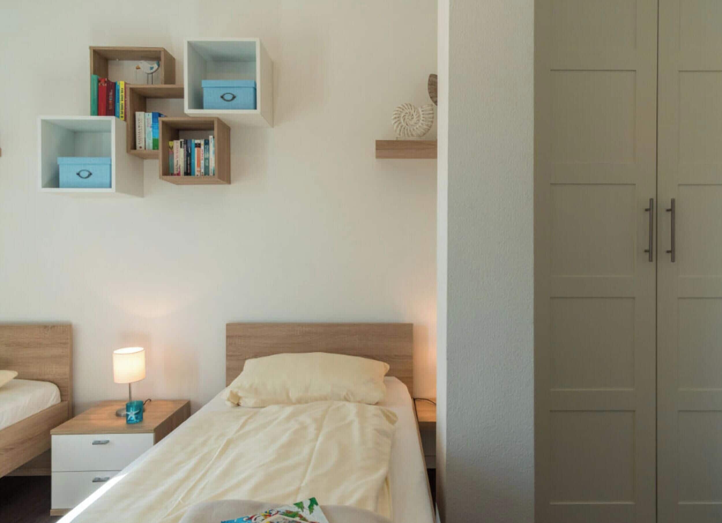 Quarto com cama, mesas de cabeceira, abajur e estantes embutidas com livros e objetos decorativos.