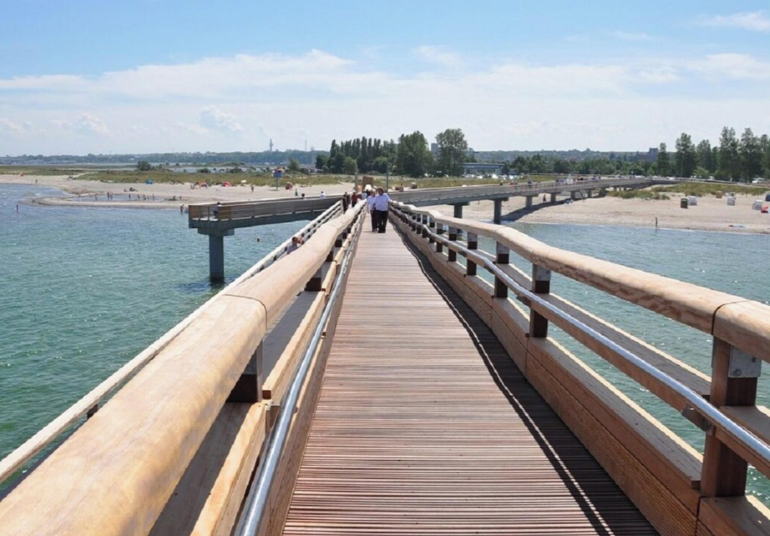 Ein hölzerner Steg führt zu einem Strand mit freiem Blick auf das Wasser und entfernte Gebäude.
