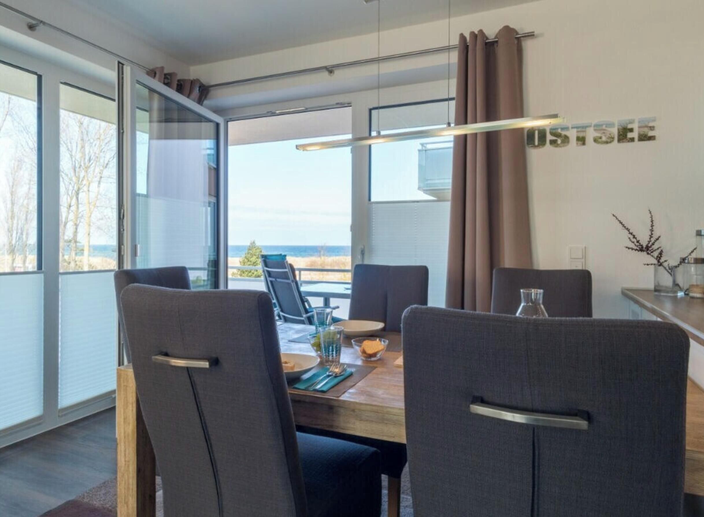 A modern dining area with a view of the sea, featuring a wooden table, grey upholstered chairs, and a pendant light.