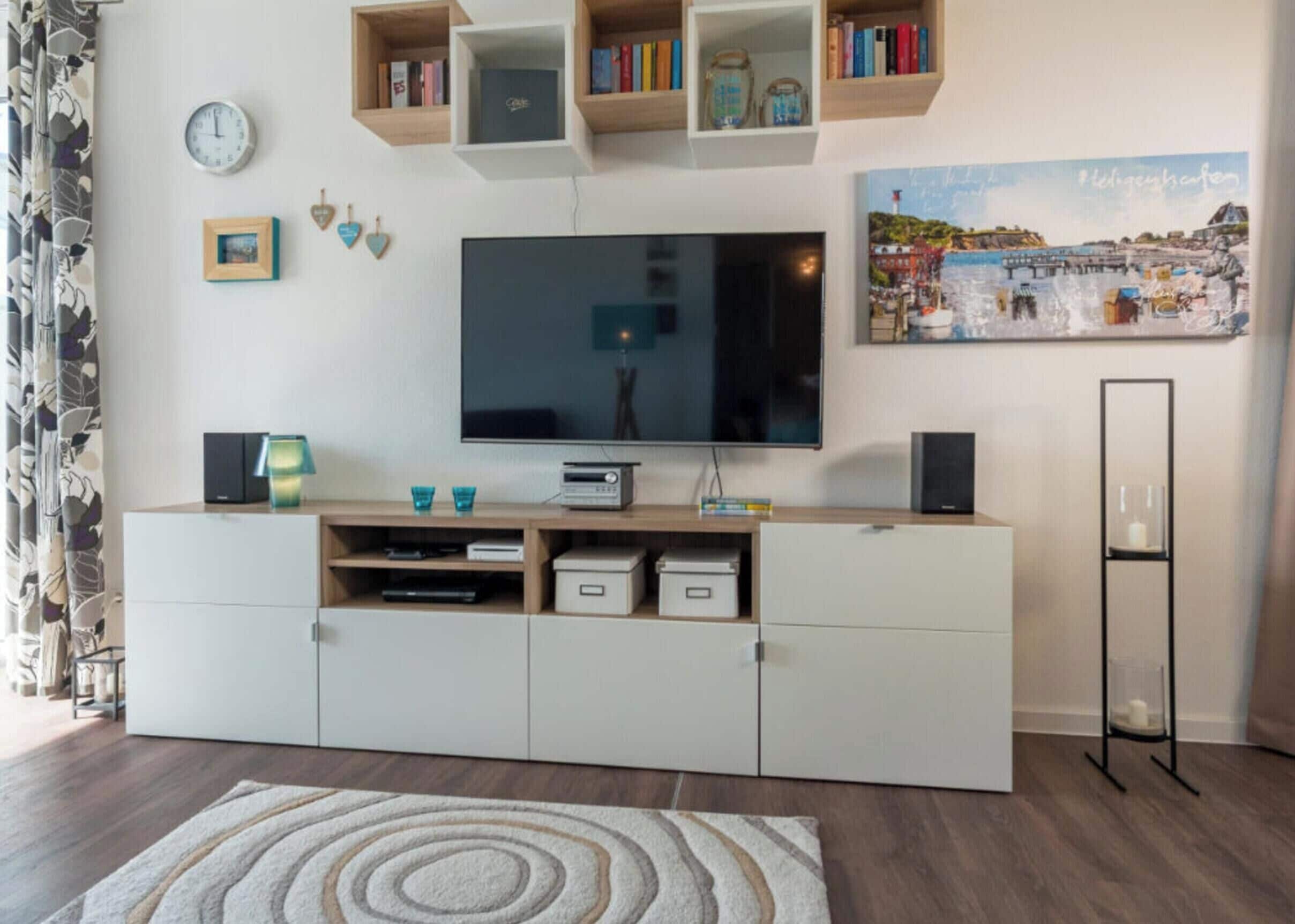Una sala de estar con un televisor de pantalla plana, estanterías de madera con libros, un reloj y un cuadro enmarcado.
