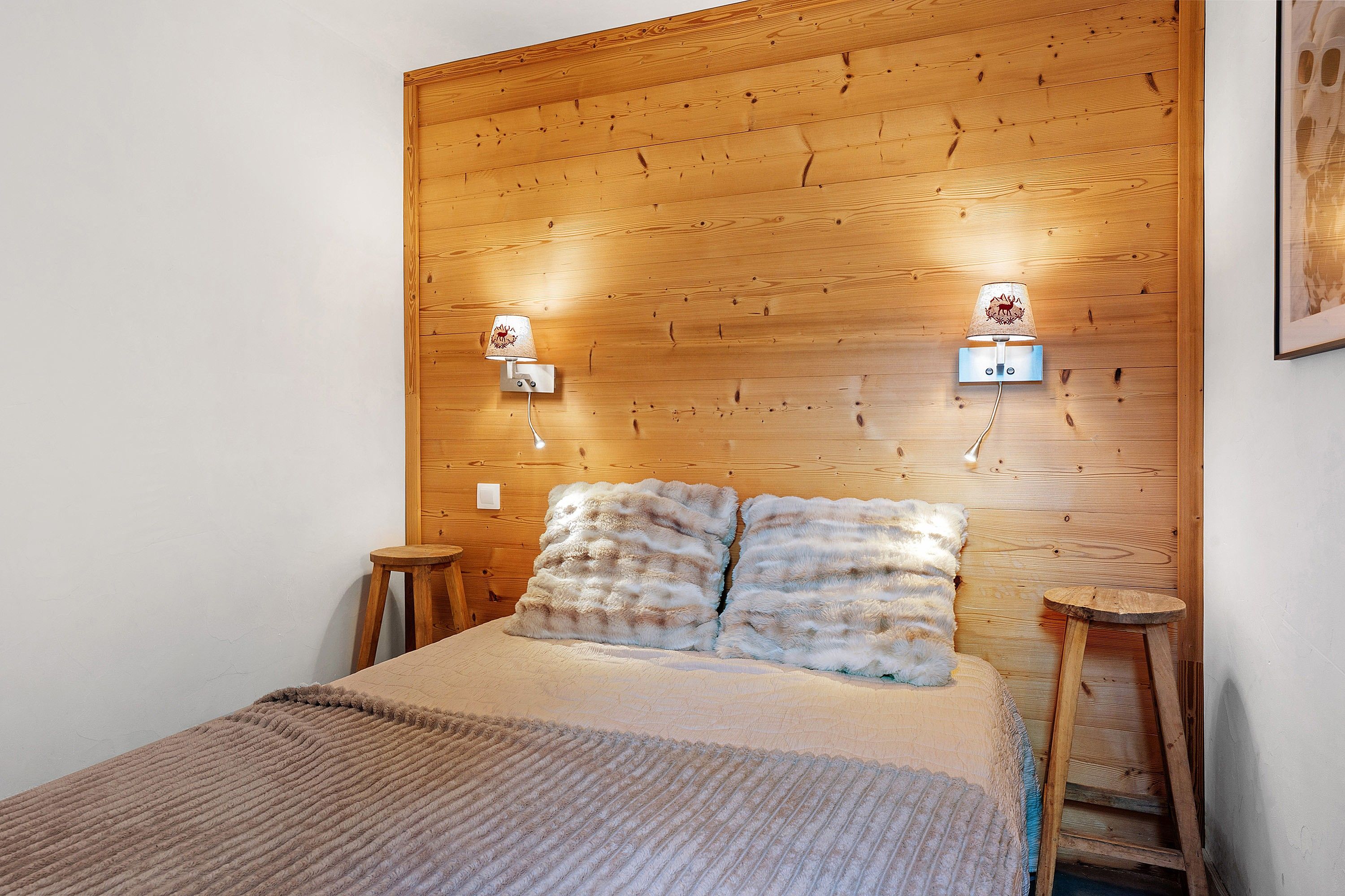 Een slaapkamer met een houten hoofdeinde, twee wandlampen, een bed met een zachte deken en twee houten krukjes.