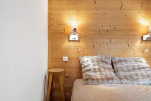 A room with a wooden wall, a bed with fluffy pillows, a small wooden stool, and a wall-mounted light fixture.