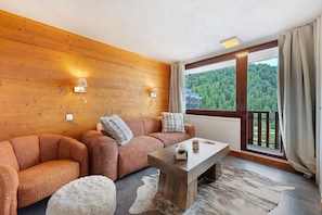 A modern living room with a wooden wall, a sofa set, a coffee table, and a view of a forested area through a large window.