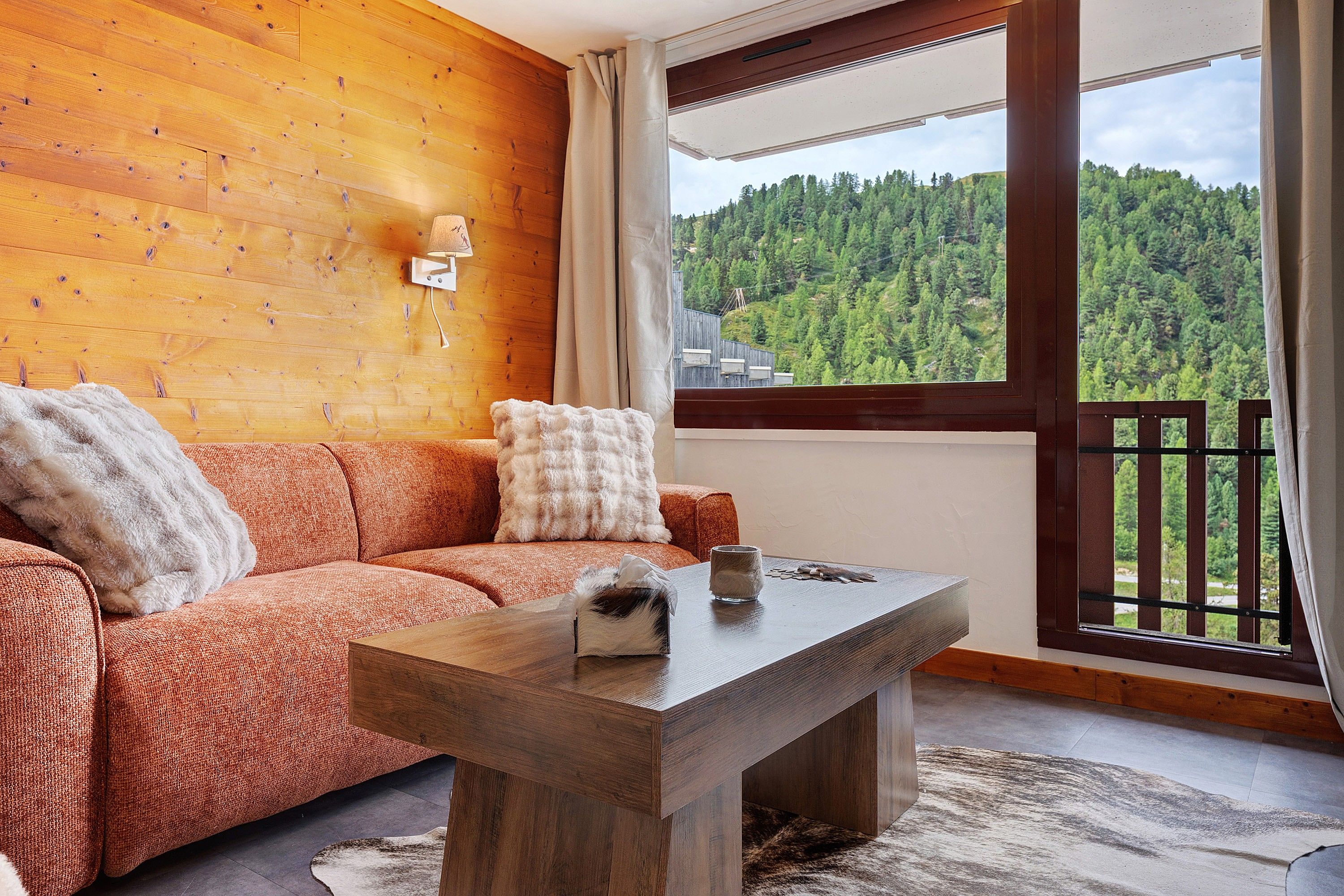 A cozy room with a wooden wall, a sofa with a fluffy pillow, a wooden coffee table, and a view of a forested hillside through a window.