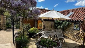 Une terrasse avec une table et des chaises sous un parasol blanc, entourée de plantes en pot et d’un mur de pierre.