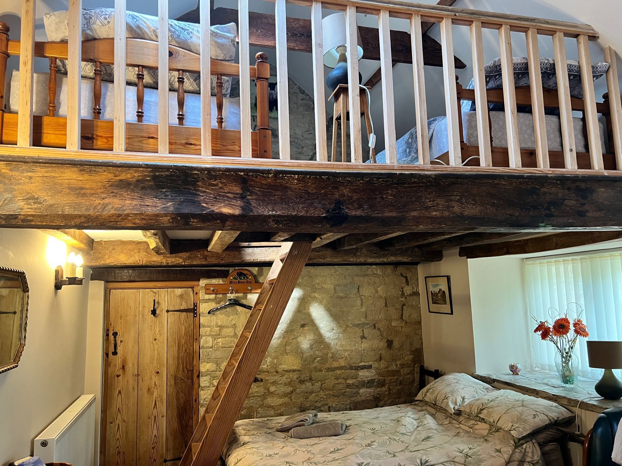 A loft bedroom with a wooden staircase, a bed with floral bedding, a stone wall, and a window with flowers.