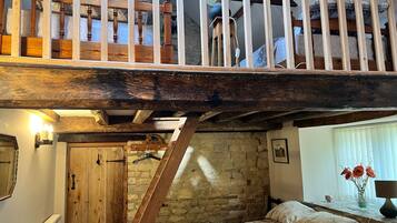 A loft bedroom with a wooden staircase, a bed with floral bedding, a stone wall, and a window with flowers.