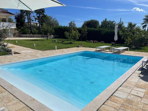 A rectangular swimming pool with a stone border, surrounded by a patio area with lounge chairs and a white umbrella. - Detached Villa for Exclusive Use with an Infinity Pool (Puglia)