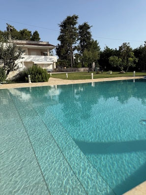 A clear swimming pool with a tiled edge, surrounded by a grassy area and a building with a stone facade. - Detached Villa for Exclusive Use with an Infinity Pool (Puglia)