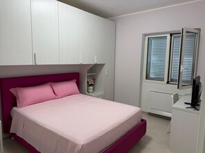 Quarto com cama, escrivaninha, janela com persianas e guarda-roupa branco.