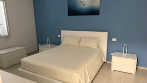 Ett sovrum med en stor säng, två sängbord, ett litet bord, en bänk och en tavla på väggen.