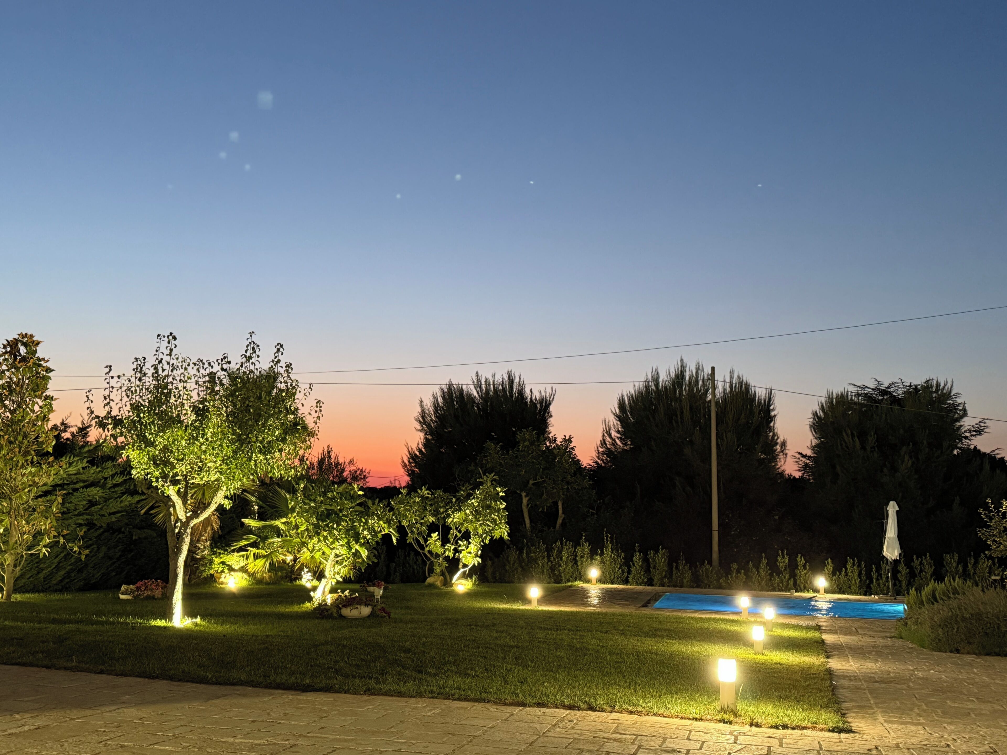 A nighttime outdoor area with a swimming pool, illuminated by multiple lights, surrounded by trees and a clear sky.