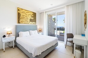 Ein Hotelzimmer mit einem Bett, einem Schreibtisch, einem Stuhl, einem Nachttisch, einer Lampe und einem großen Fenster mit Blick auf das Meer.