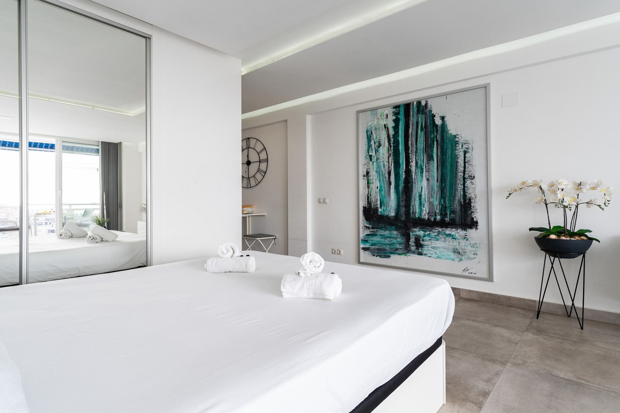 Une chambre d’hôtel moderne avec un grand lit, un tableau, une orchidée en pot et une horloge au mur.