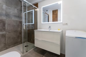 Ett modernt badrum med en duschkabin i glas, ett vitt handfat med en spegel ovanför och en vit tvättmaskin.