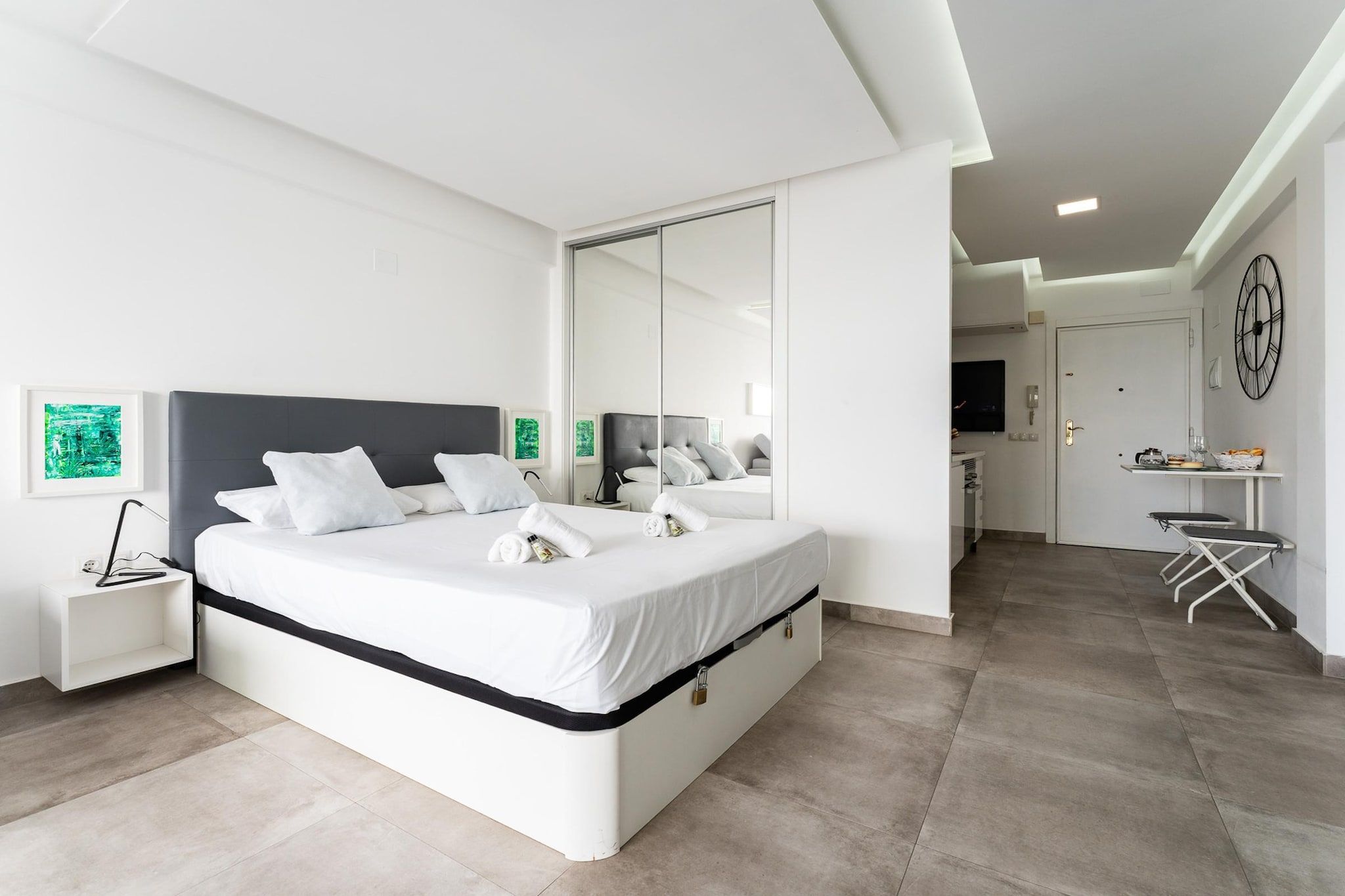 Ein modernes Schlafzimmer mit einem großen Bett, einem Nachttisch, einem Spiegel und Blick in den angrenzenden Wohnbereich.