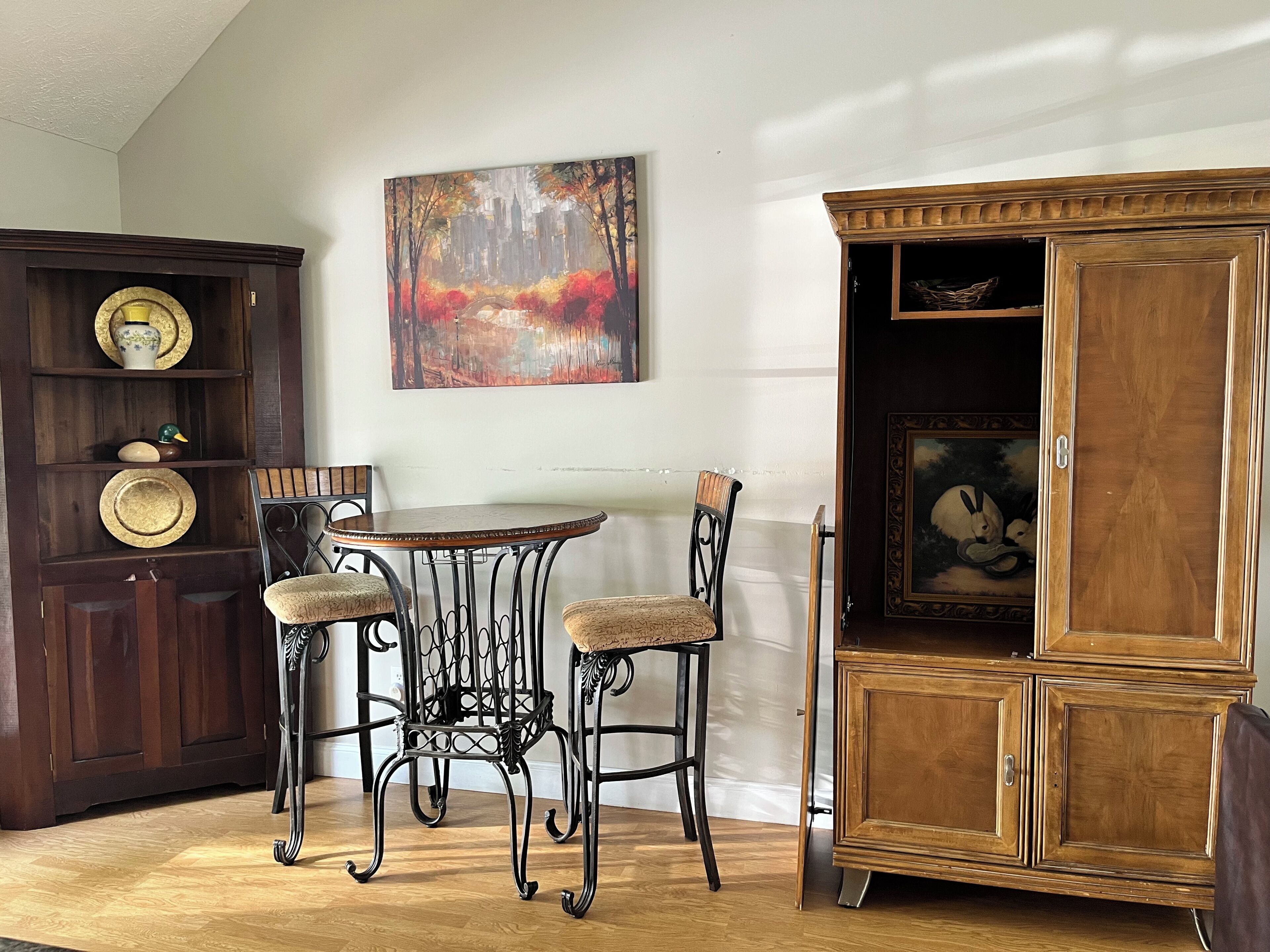 A dining area with a round table and two chairs, a wooden cabinet with a painting, and a shelf with decorative items.