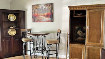 A dining area with a round table and two chairs, a wooden cabinet with a painting, and a shelf with decorative items.