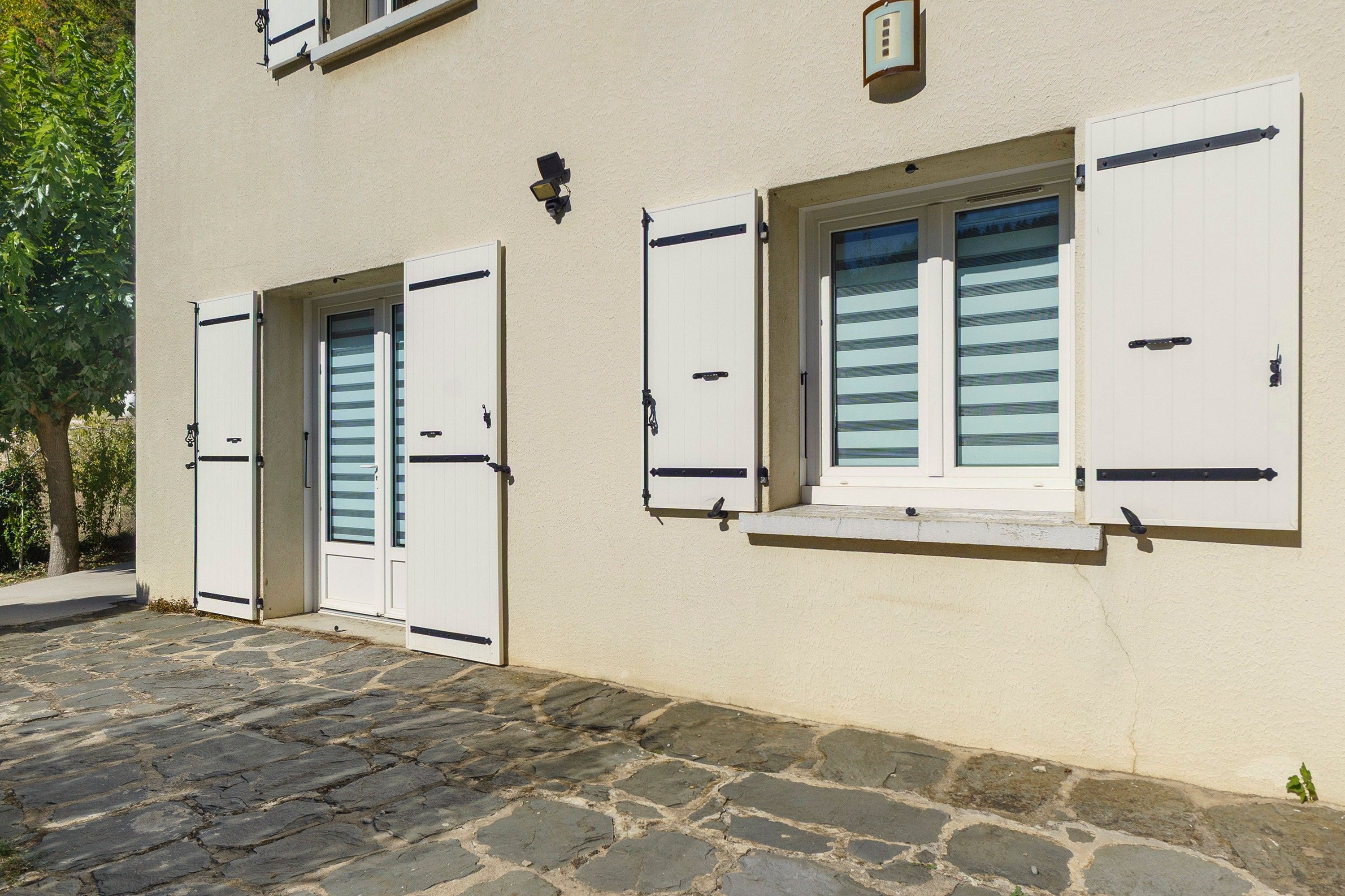 Un bâtiment doté de deux portes blanches et de fenêtres avec volets, une cour pavée de pierres et un mur à la texture particulière.