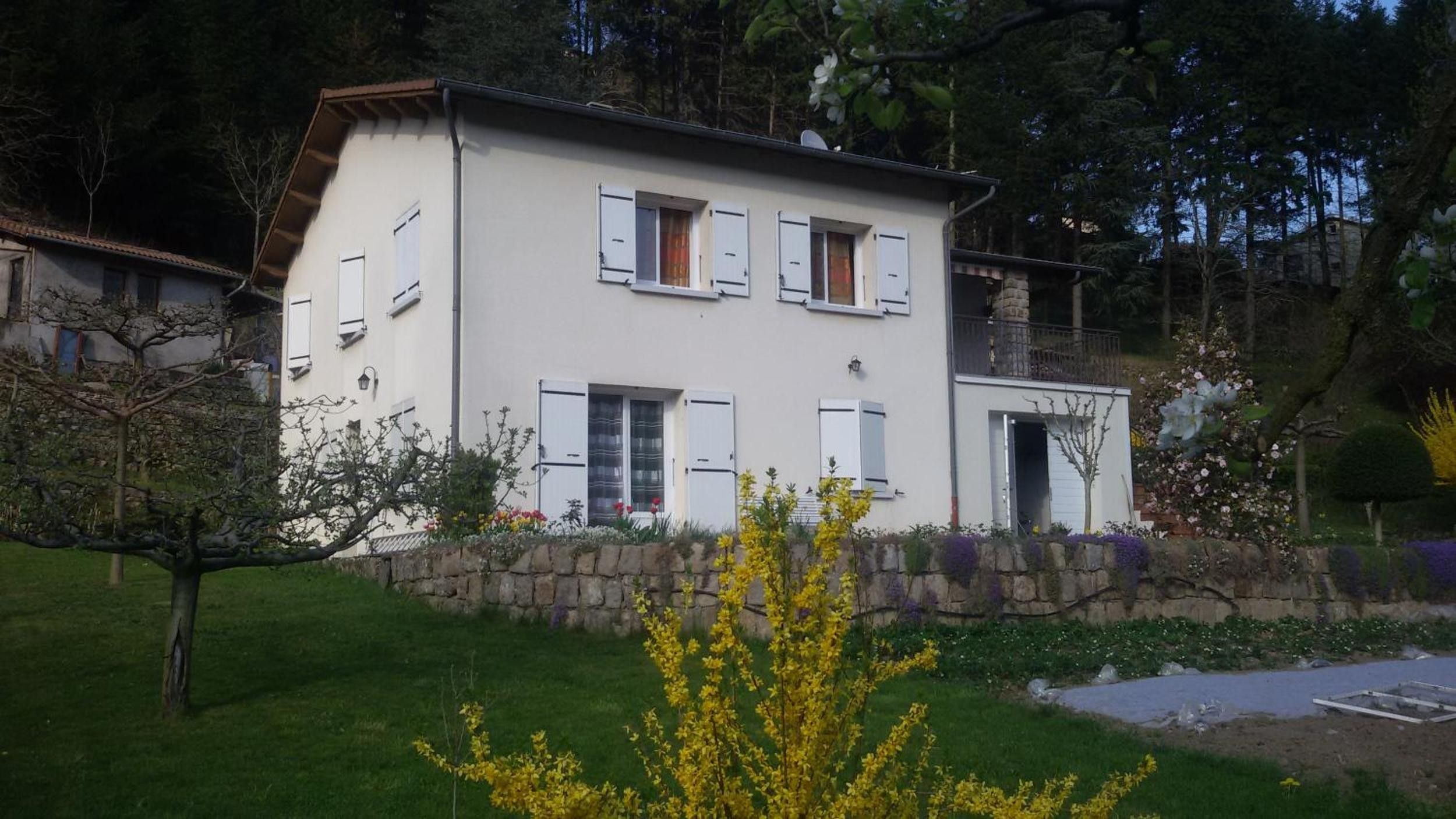 Une maison à deux étages, avec un mur de pierre à l’avant, entourée de verdure et d’arbres.