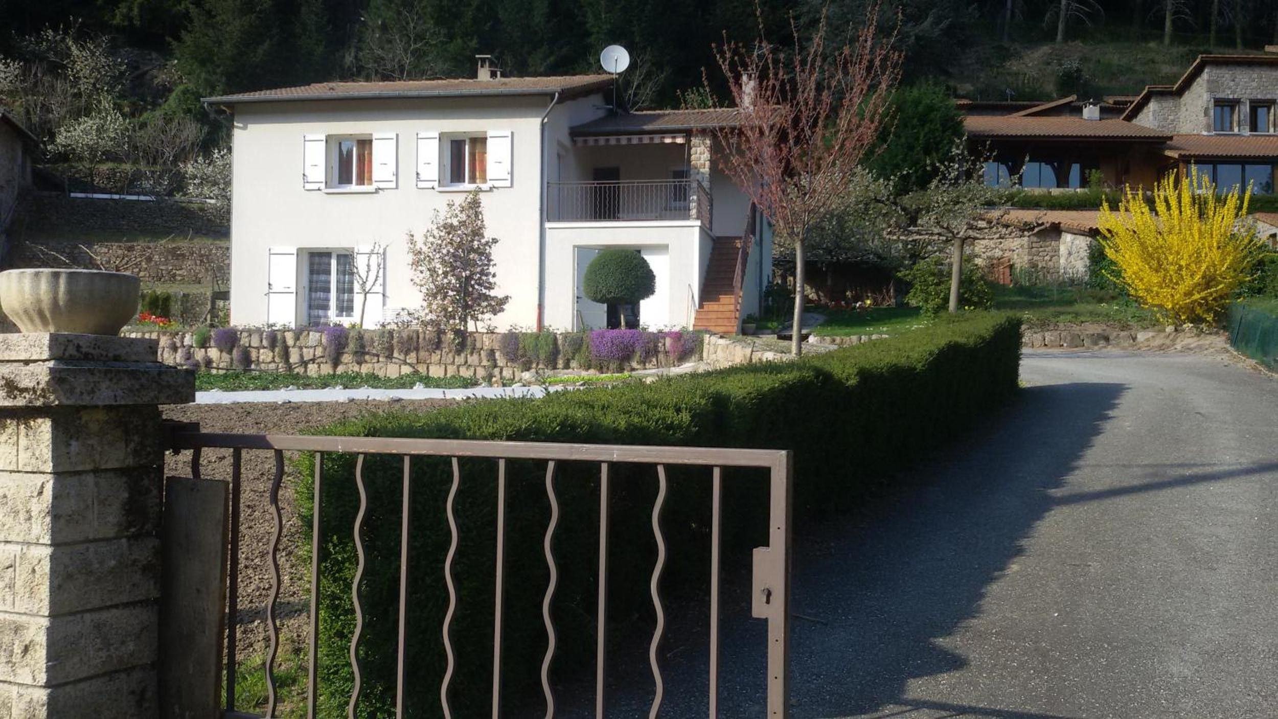 Una casa de dos pisos con entrada protegida por un portón, un pequeño jardín con diversas plantas y un cielo despejado.