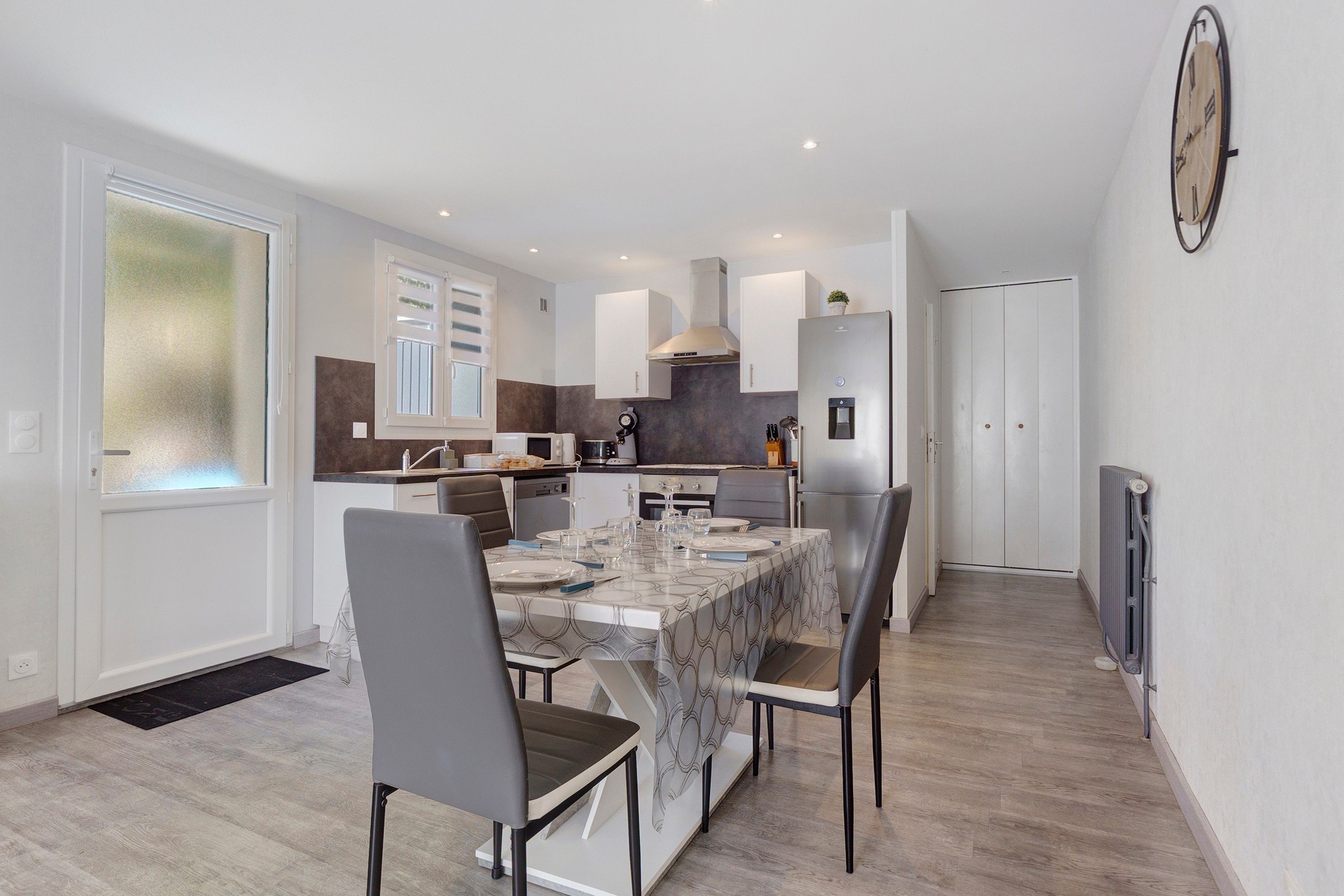 A modern kitchen with a dining area, equipped with stainless steel appliances and a large clock on the wall.