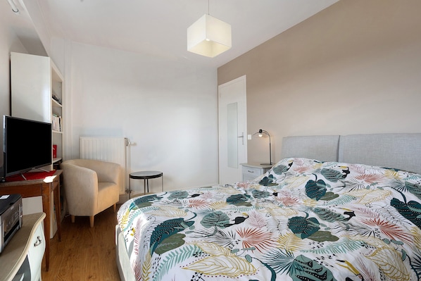 A bedroom with a bed, a chair, a desk with a computer, a television, and a bedside lamp.