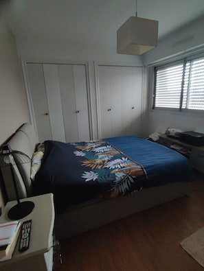 Un dormitorio con cama, escritorio, silla, lámpara, ventana con persianas y armario.