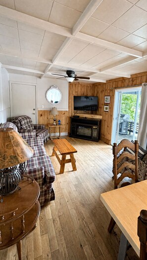 Ein gemütliches Wohnzimmer mit einer Sofagarnitur, einem Holztisch, einem an der Wand montierten Fernseher und einem Deckenventilator.