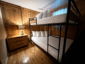 Ein Schlafraum mit Etagenbetten, Holzboden, einem Nachttisch mit Lampe und einem Fenster mit Spitzevorhängen.