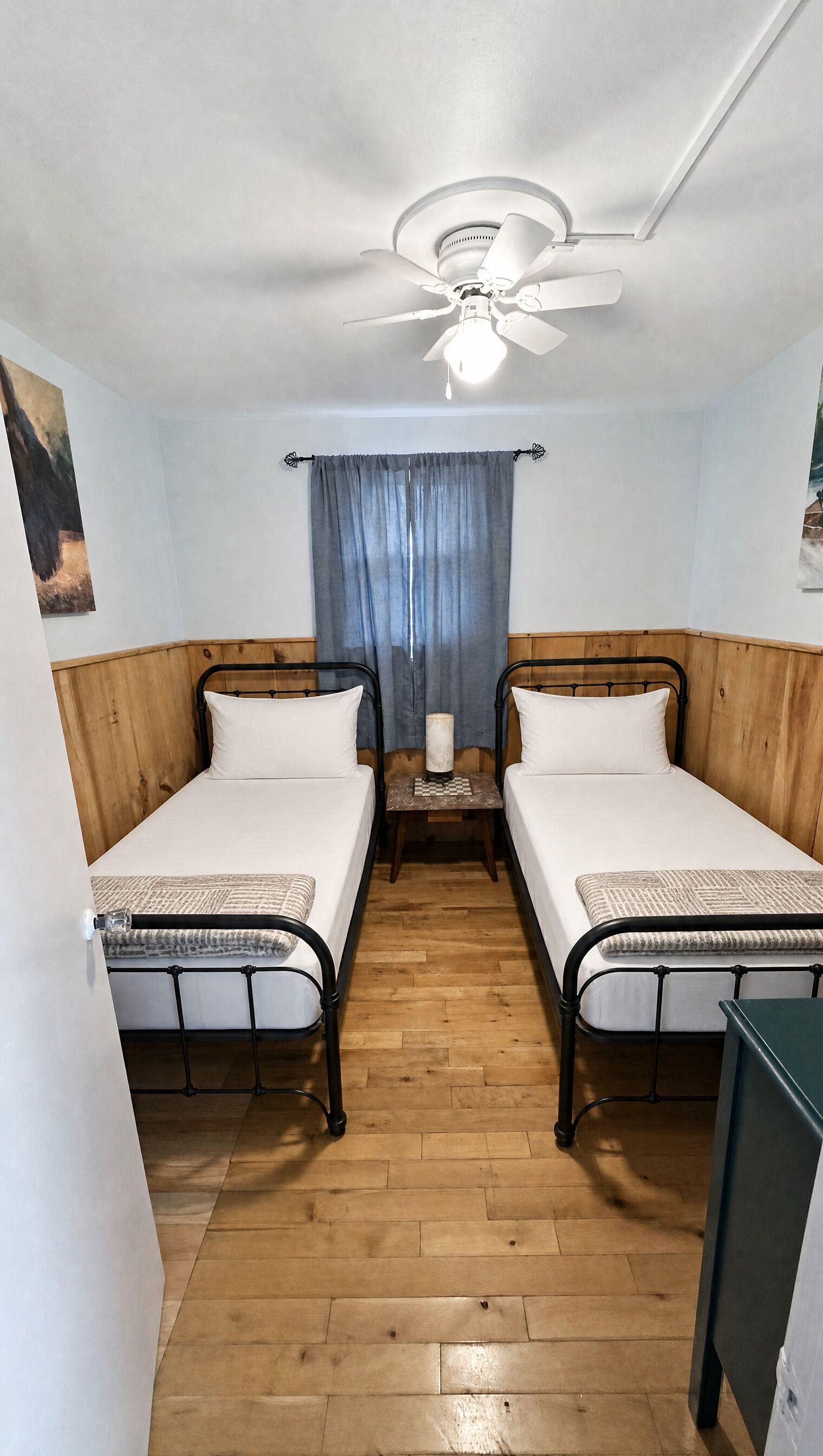A room with two single beds, a ceiling fan, a small table with a lamp, and a wooden floor.