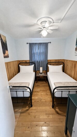 Ein Zimmer mit zwei Einzelbetten, einem Deckenventilator, einem kleinen Tisch mit Lampe und Holzboden.