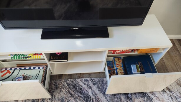 Una TV a schermo piatto su un mobile bianco contenente scaffali con giochi da tavolo e libri.