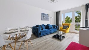 Una sala moderna con un sofá azul, un sillón amarillo, una mesa de comedor preparada para cuatro personas y un ventanal con cortinas.
