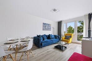 Una sala moderna con un sofá azul, un sillón amarillo, una mesa de comedor preparada para cuatro personas y un ventanal con cortinas.