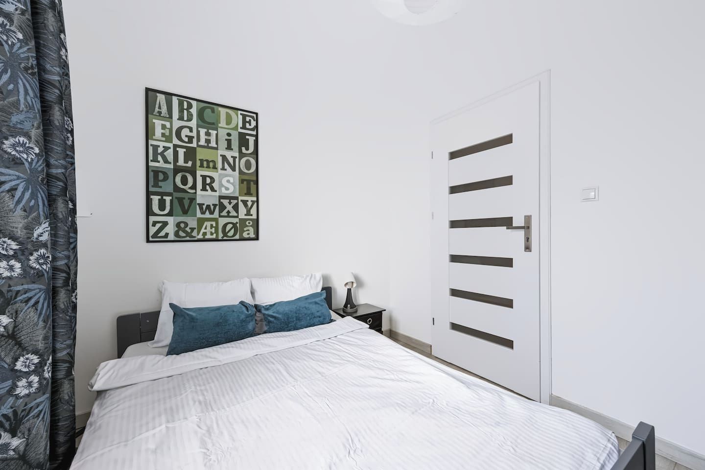 Ein Schlafzimmer mit Bett, Kissen, einem Nachttisch, einem an der Wand hängenden Alphabet-Poster und einer modern gestalteten Tür.